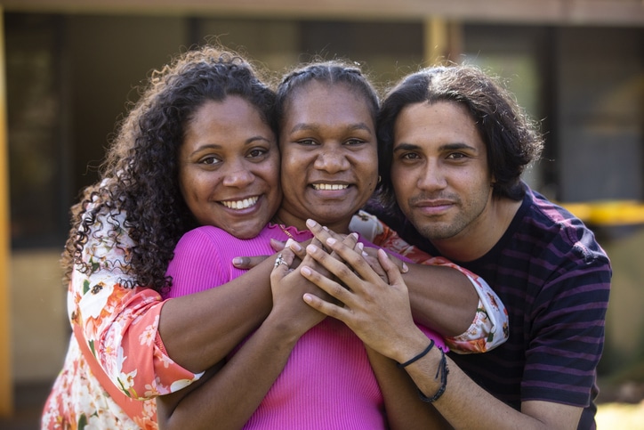 Three young aboriginal students outdoors with their arms around each other smiling at the camera.