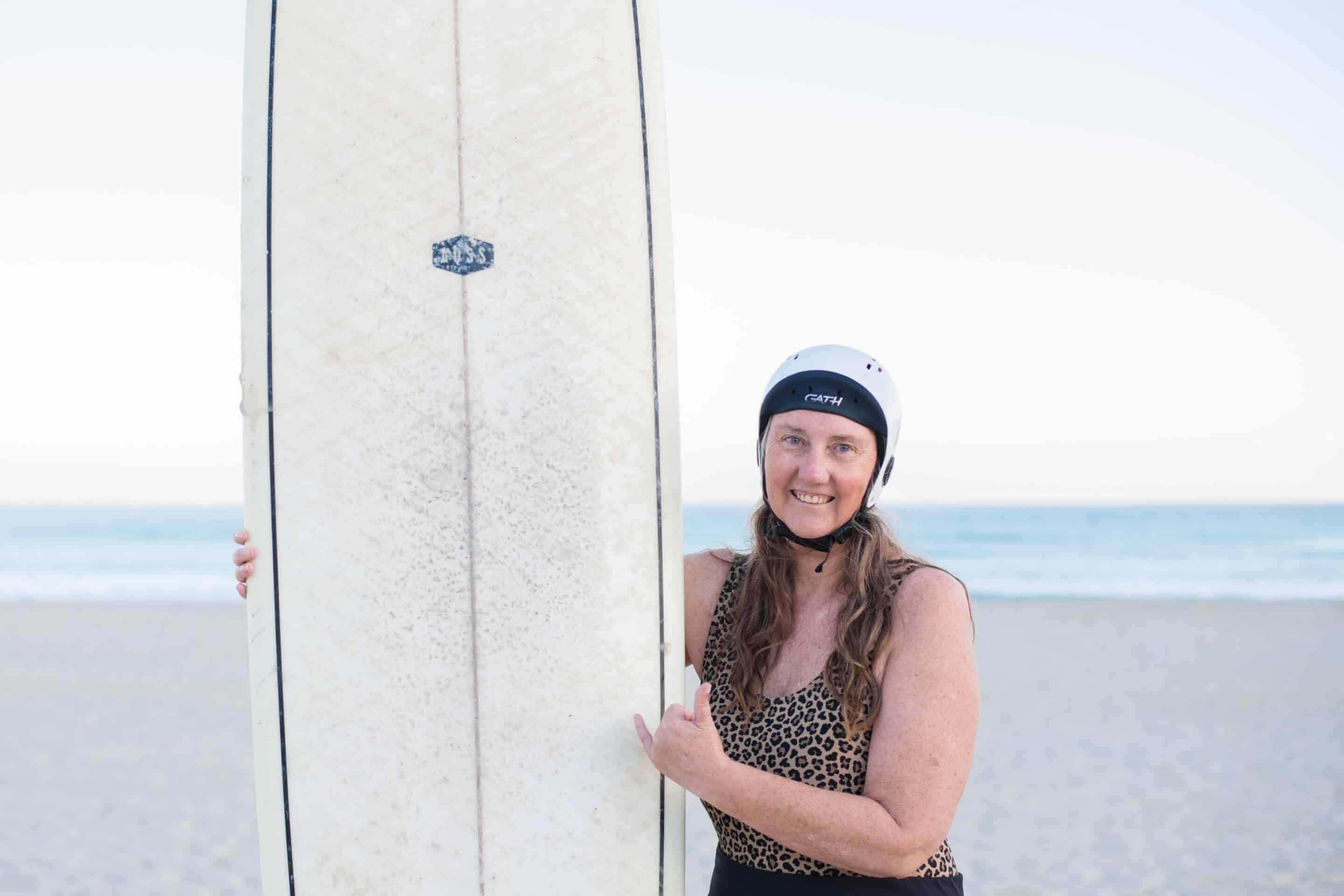 photo of a woman on the beach wearing her surfing helmet and holding a long-board 