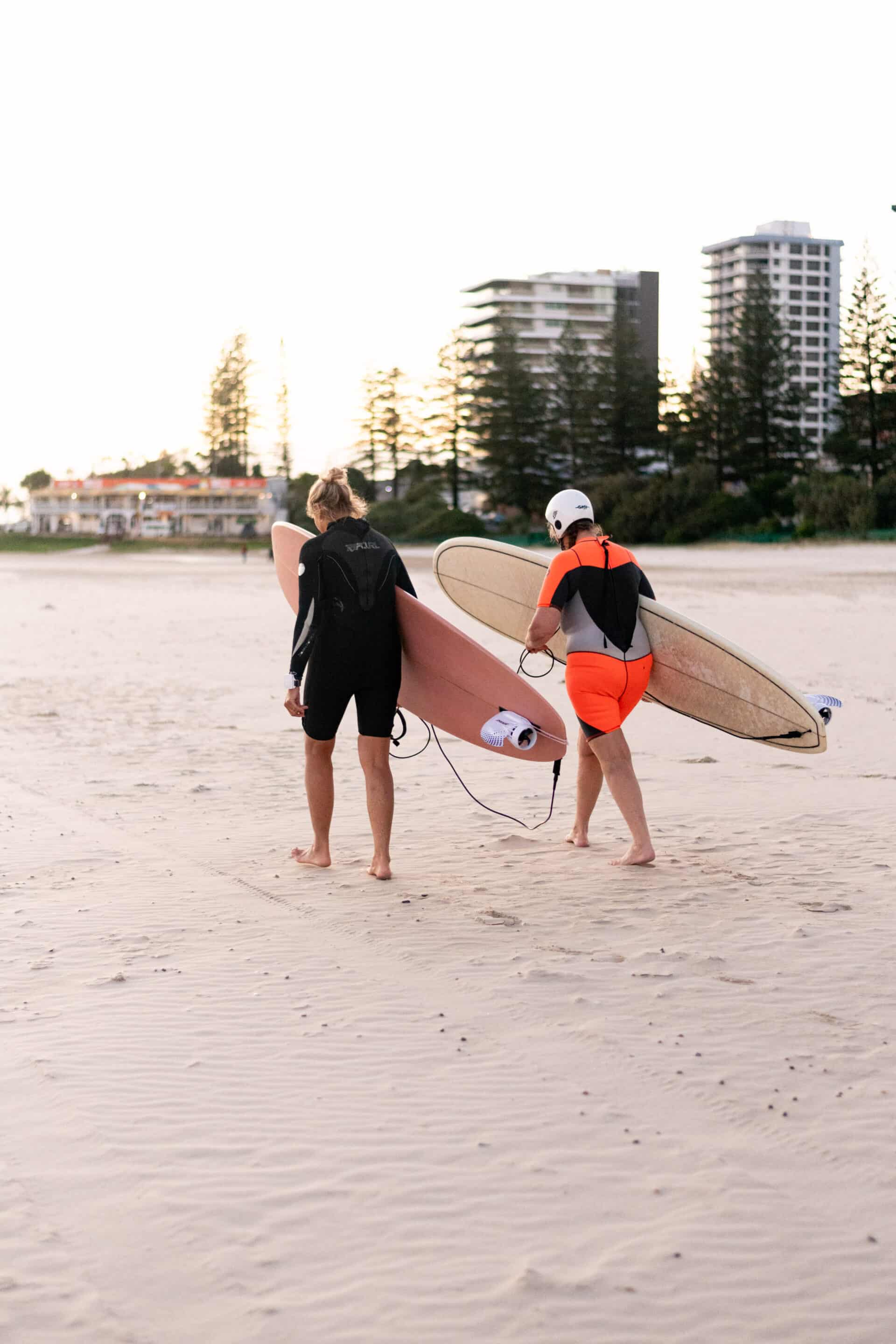 2 people standing on beach with surfboards in arms, wearing wetsuits
