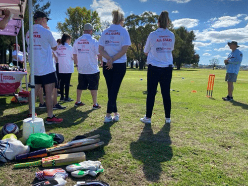 Shows Scott teaching the Community All Stars the rules of the game - with cricket bats and gloves in the foreground
