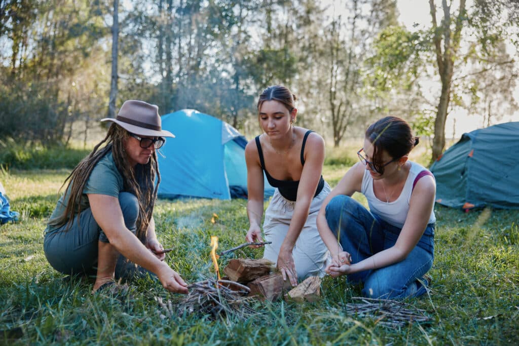 Photo posed by models: Three women lighting a fire camping. 