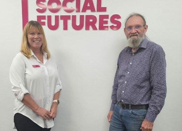 Linda Hayes and Dr Gregory Smith stand beside each other smiling in front of a magenta Social Futures sign