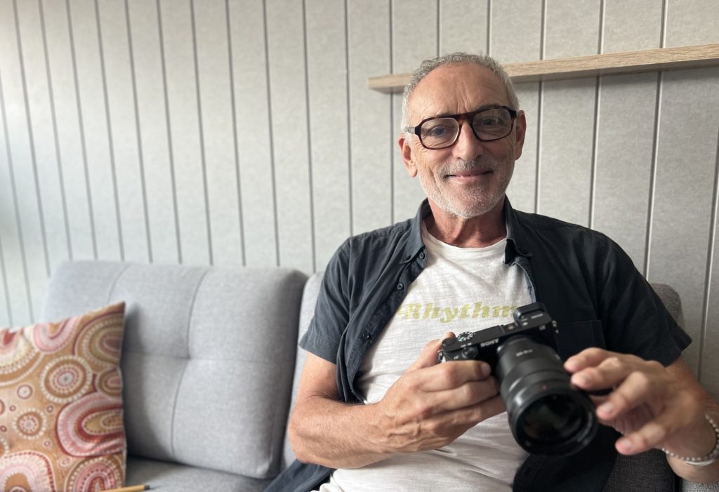 Vinnie wears glasses and holds a camera, he is sitting on a couch smiling