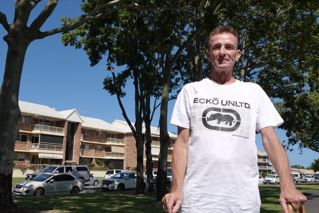 Richard wears a white t-shirt and stands near his apartment in Tweed Heads